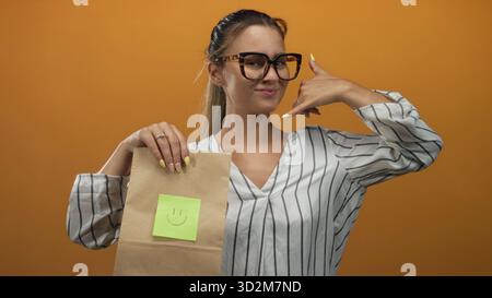 Frau, die eine Brille trägt, eine Papiertüte mit einem lächelnden Haftnotiz hält und mit dem Finger ins Studio zeigt; freundliches Vertrauen. Stockfoto