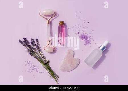 Ein Set von Kosmetikprodukten in Glasflaschen, ein Massagegerät und ein Rosenquarzschaber, frischer Lavendelzweig. Flache Lagen. Draufsicht. Home Spa Care Stockfoto