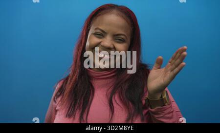 afroamerikanerin winkt Hand im Studio, trägt einen rosa Rollkragen und lächelt warm, während sie eine Smartwatch trägt; Gruß. Stockfoto