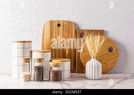 Set aus weißen Keramikgläsern, Gewürzgläsern aus Glas, Schneidebrettern aus Holz auf Marmorarbeitsfläche. Küchenhintergrund Stockfoto