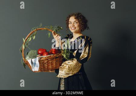 Porträt einer jungen erwachsenen Frau in einem mittelalterlichen Kleid, die einen Korb mit Gemüse und Früchten in der Hand hält Stockfoto