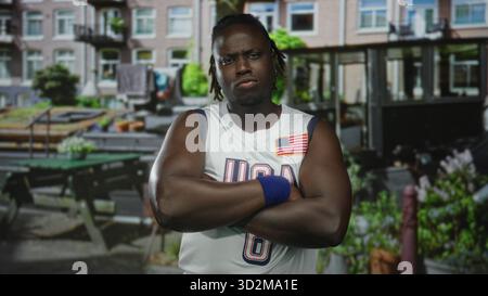 Die Arme des Mannes kreuzten sich, tragend ein us-Basketballtrikot und blaues Armband, Flaggenaufnäher sichtbar, stehend in einem städtischen Straßencafé mit Bänken und Pflanzgefäßen; def Stockfoto