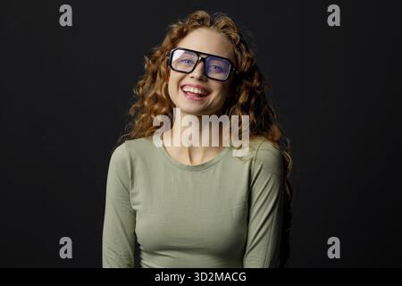 Glückliche lächelnde, lockige junge Frau, die eine Brille trägt und mit einem fröhlichen Lächeln auf schwarzem Hintergrund in die Kamera schaut Stockfoto
