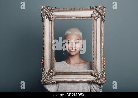 Eine junge Frau mit einem Lächeln und kurzen blonden Haaren hält einen vergoldeten Bilderrahmen in den Händen vor ihrem Gesicht Stockfoto