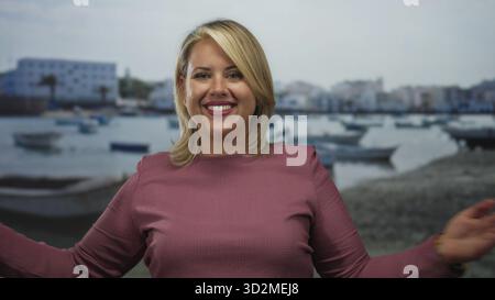 Frau streckt die Arme in offener Umarmung am sonnendurchfluteten Sandstrand, während sie breit lächelt; Freude willkommen. Stockfoto