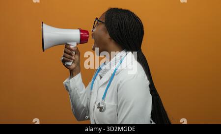 Frau Ärztin mit Stethoskop, die durch Megaphon vor orangem Hintergrund schreit, Begeisterung und Kommunikation ausdrückt. Stockfoto