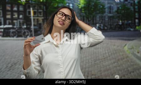 Frau, die die Kreditkarte in der Nähe der Kamera hält, während sie mit freier Hand auf einer kopfsteingepflasterten Straße in der Stadt Gesten macht, Brille trägt; Zahlungsfrustration. Stockfoto