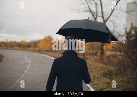 Junger Mann im schwarzen Mantel wandern im Regen mit Schirm Stockfoto