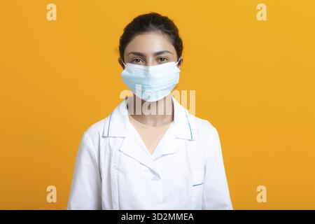Porträt der indischen Ärztin, die weißes medizinisches Kleid und Gesichtsmaske trägt, isoliert auf gelbem Hintergrund Stockfoto