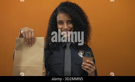 Junge hispanische Polizistin, die Kaffee- und Papiertüte in Uniform vor orangem Hintergrund hält und selbstbewusst lächelt. Stockfoto