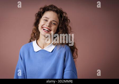 Glückliche lächelnde junge Erwachsene Frau trägt einen winterblauen Pullover drinnen und schaut mit freudigem Lächeln auf die Kamera Stockfoto