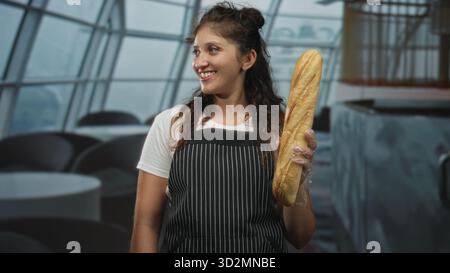 Frau, die Baguette mit Handschuhen im Restaurant-Essbereich des Gebäudes hält, gestreifte Schürze trägt; herzliche Gastfreundschaft. Stockfoto