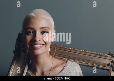 Die junge Frau mit ihren kurzen blonden Haaren hält einen vergoldeten Bilderrahmen in den Händen auf ihrem Nacken Stockfoto