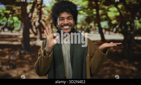 Lächelnder Mann im Park, der Handgeste mit Bäumen im Freien in grünem Hintergrund zeigt und Schal und Mantel trägt. Stockfoto