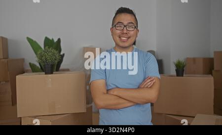 Junger chinesischer Mann mit Armen, die neben gestapelten Pappschachteln im Gebäude während des Umzugs gekreuzt wurden; Zufriedenheit des beweglichen Tages. Stockfoto