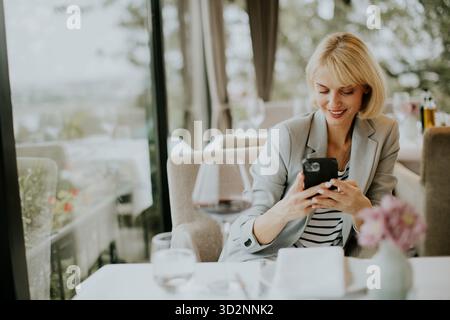 Eine Frau sitzt bequem in einem Café und lächelt, während sie ihr Smartphone benutzt. Sonnenlicht filtert durch große Fenster und schafft eine warme Atmosphäre. Tabellen sind Stockfoto