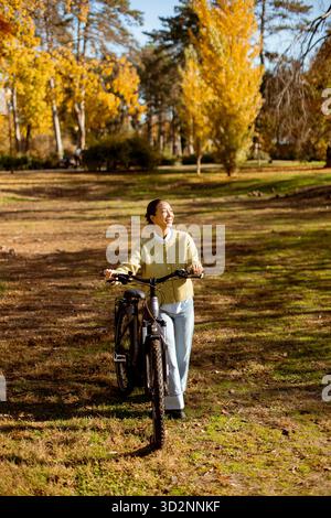 Eine junge Frau läuft mit dem Fahrrad auf einem ruhigen Pfad, umgeben von lebhaften Herbstbäumen und goldenen Blättern, und genießt die warme Sonne. Stockfoto