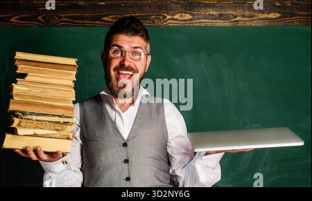 Männlicher Schüler mit Büchern und Laptop vor der Tafel im Klassenzimmer. Der Universitätslehrer hält Lehrbücher und Laptops. Lächelnder Student in gla Stockfoto