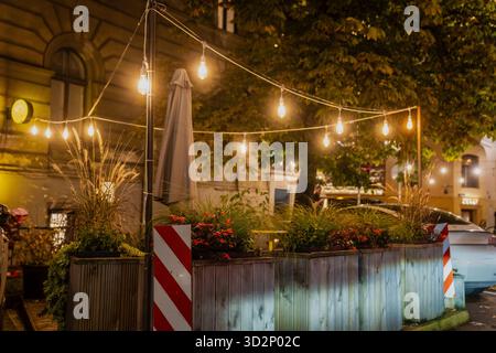 Urbane Außenterrasse mit dekorativen Lichtern, die Holzpflanzen mit ornamentalen Gräsern und Blumen beleuchten und eine gemütliche Abendstimmung schaffen Stockfoto