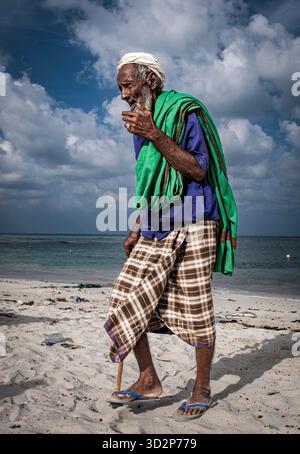 Ein Fischer spaziert entlang des Sandstrandes in Qulensya, Socotra, und genießt einen ruhigen Moment in der Nähe des Meeres unter einem teilweise bewölkten Himmel. Stockfoto