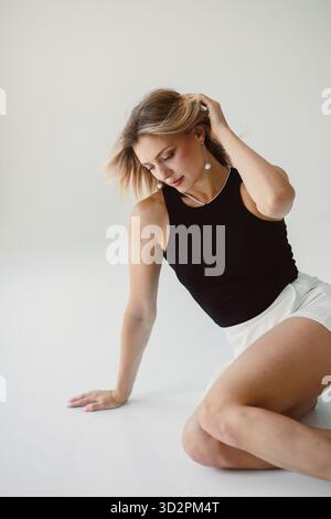Eine junge Frau sitzt anmutig auf einem glatten Boden in einem hellen Studio. Sie trägt ein schwarzes Oberteil und weiße Shorts und strahlt Eleganz aus, während sie Witz spielt Stockfoto