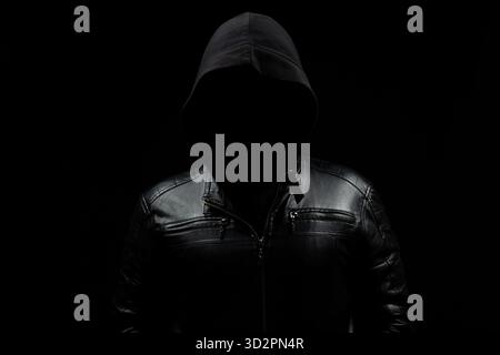 Nahaufnahme eines gesichtslosen Hacker-Mannes mit Kapuze im Schatten mit Lederjacke und schwarzem Hintergrund Stockfoto