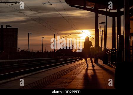 Die Silhouette einer Person, die während eines dramatischen Sonnenuntergangs auf einem Bahnsteig steht, lange Schatten und Überlandleitungen schaffen eine stimmungsvolle Atmosphäre Stockfoto