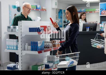 Weibliche Klientin überprüft Etiketten Informationen zu Vitaminen, die in Apothekenregalen, Einzelhandelsservice in einem Gesundheitsbereich platziert werden. Asiatische Frau auf der Suche nach Wellness-Ergänzungen, Apotheke Besuch. Stockfoto