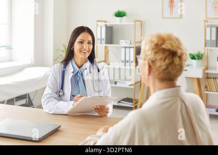 Lächelnde Kardiologin mit Klemmbrett, Gespräch mit älteren Patienten in hellem Arztpraxis. Stockfoto