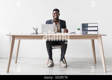 Junger schwarzer Geschäftsmann, der Kaffee genießt, während er online an seinem Schreibtisch in einem Studio arbeitet Stockfoto