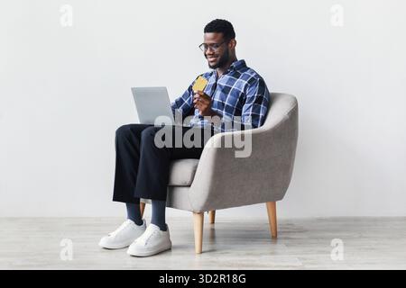 Junger Mann genießt Online-Shopping mit Kreditkarte und Laptop in modernem Stuhl Stockfoto