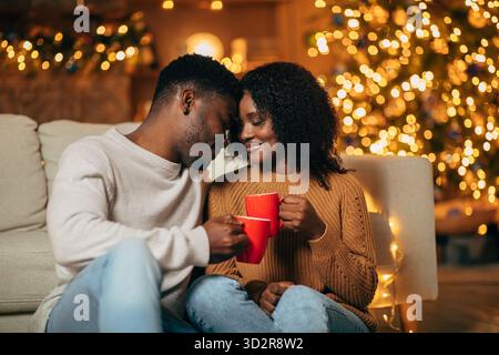 Wunderschönes schwarzes Paar teilt sich einen liebevollen Moment mit Kaffee am Heiligabend in einem gemütlichen, dekorierten Zimmer Stockfoto