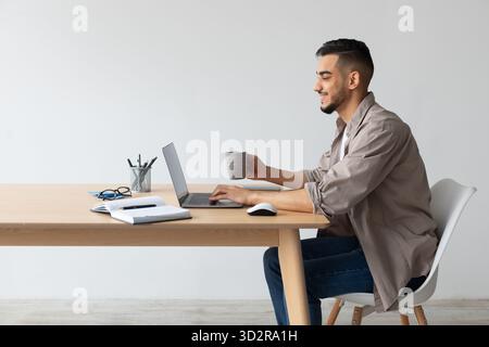 Lächelnder arabischer Mann zu Hause mit Laptop und Kaffee beim Arbeiten am Schreibtisch Stockfoto