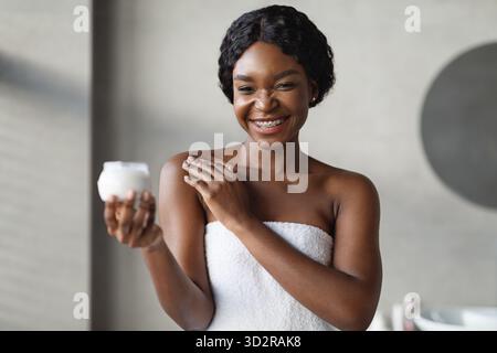 Lächelnde junge schwarze Frau, die nach der Dusche Körpercreme in einem modernen Badezimmer aufträgt Stockfoto
