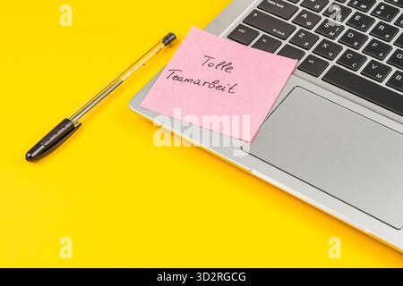 Ein Laptop sitzt auf einer hellgelben Oberfläche mit einem Haftnotiz mit der Aufschrift tolle Teamarbeit. Neben dem Laptop befindet sich ein Stift, der eine produktive Arbeit widerspiegelt Stockfoto