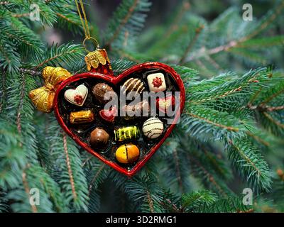 Weihnachtsschmuck in Herzform mit Pralinen im Inneren. Schokoladenbox mit Baumhintergrund. Stockfoto