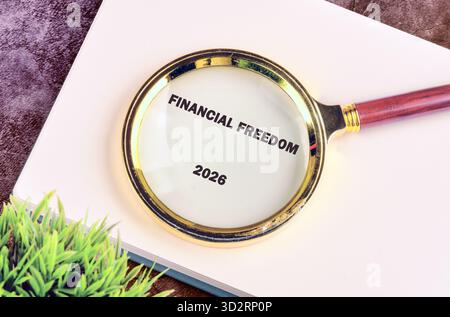 Eine Lupe konzentriert sich auf die Worte Financial Freedom 2026, die auf einer Seite geschrieben sind. Neben dem Notizblock befindet sich eine kleine grüne Pflanze, die auf Wachstum und hindeutet Stockfoto
