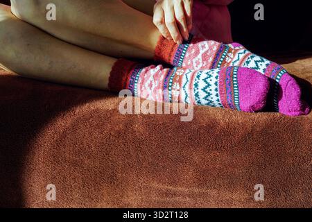 Die Füße einer Frau in skandinavischen Stricksocken liegen auf einem mit Decke bedeckten Bett. Das Konzept von Hygge-inspiriertem Komfort, entspanntem Lifestyle und wohliger Wärme Stockfoto