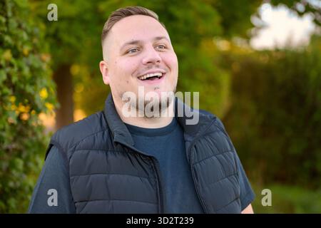 Ein fröhlicher junger Mann lacht offen, während er draußen in einer natürlichen grünen Umgebung bei Tageslicht steht. Stockfoto