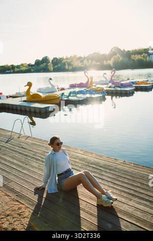Eine Frau sitzt friedlich auf dem Dock und genießt die Sonne, während lebhafte Paddelboote sanft auf dem See im Hintergrund schweben und eine ruhige und fröhliche Atmosphäre schaffen Stockfoto