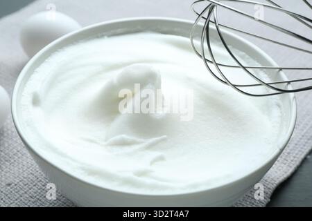 Geschlagenes weiß in Schüssel, Schneebesen und Eier auf grauem Tisch, Nahaufnahme Stockfoto