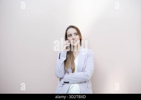 Eine Frau mit langen Haaren steht selbstbewusst und hält ein Telefon am Ohr. Sie sieht nachdenklich aus, trägt einen leichten Blazer vor einer rosa Wand. Stockfoto