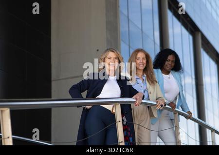 Drei Frauen mit verschiedenen Frisuren posieren auf einem stilvollen Balkon. Sie sind in modischen Outfits gekleidet, lächeln und genießen die Natur unter Stockfoto