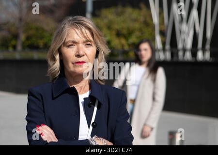 Eine selbstbewusste Frau mit blondem Haar steht mit überkreuzten Armen und trägt einen blauen Blazer. Hinter ihr läuft eine jüngere Frau in einem hellen Mantel lässig. Die sc Stockfoto