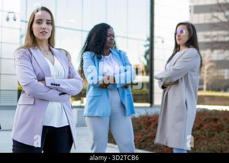 Drei Frauen mit unterschiedlichen Stilen posieren selbstbewusst außerhalb eines eleganten Büroaufbaus. Die Sonne scheint hell, wenn sie Kraft und Kameradschaft in Th zeigen Stockfoto