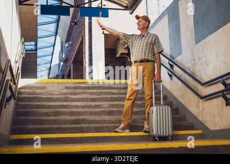Glücklicher Seniorenmann steht am Bahnhof und macht Selfie mit dem Smartphone. Senioren reisen. Stockfoto