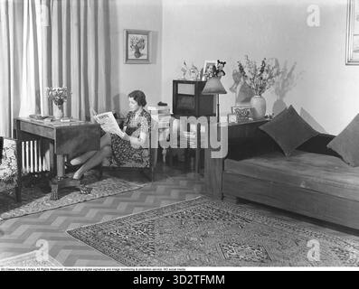 Heimleben 1941. Eine Frau liest die Tageszeitung Falbygdens Tidning in einem bescheiden eingerichteten Zimmer. Das Zimmer verfügt über ein Bett, ein Radio, zwei Sessel und einen Tisch. Teppiche bedecken den Boden. Kristoffersson Ref. 220 Stockfoto