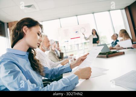 Junge Fachkräfte in einem Büro, die Dokumente während eines Geschäftsgesprächs diskutieren, während ein Teammitglied auf einem Bildschirm präsentiert Stockfoto