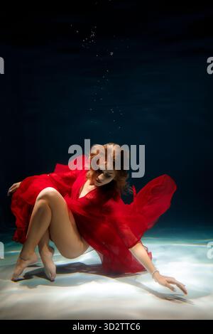 Frau in rotem Kleid posiert anmutig unter Wasser in einer ruhigen blauen Umgebung Stockfoto
