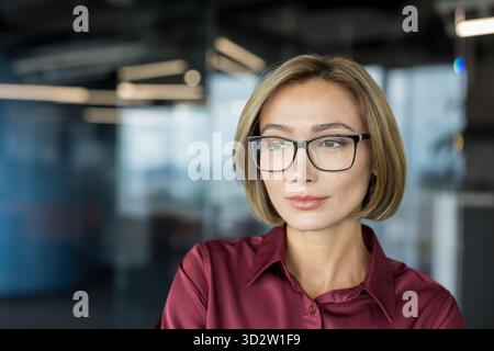 Professionelle Geschäftsfrau, die eine Brille und ein kastanienbraunes Hemd trägt, blickt selbstbewusst in eine moderne Büroumgebung und steht für Führung, Vision und anspruchsvollen Geschäftserfolg Stockfoto